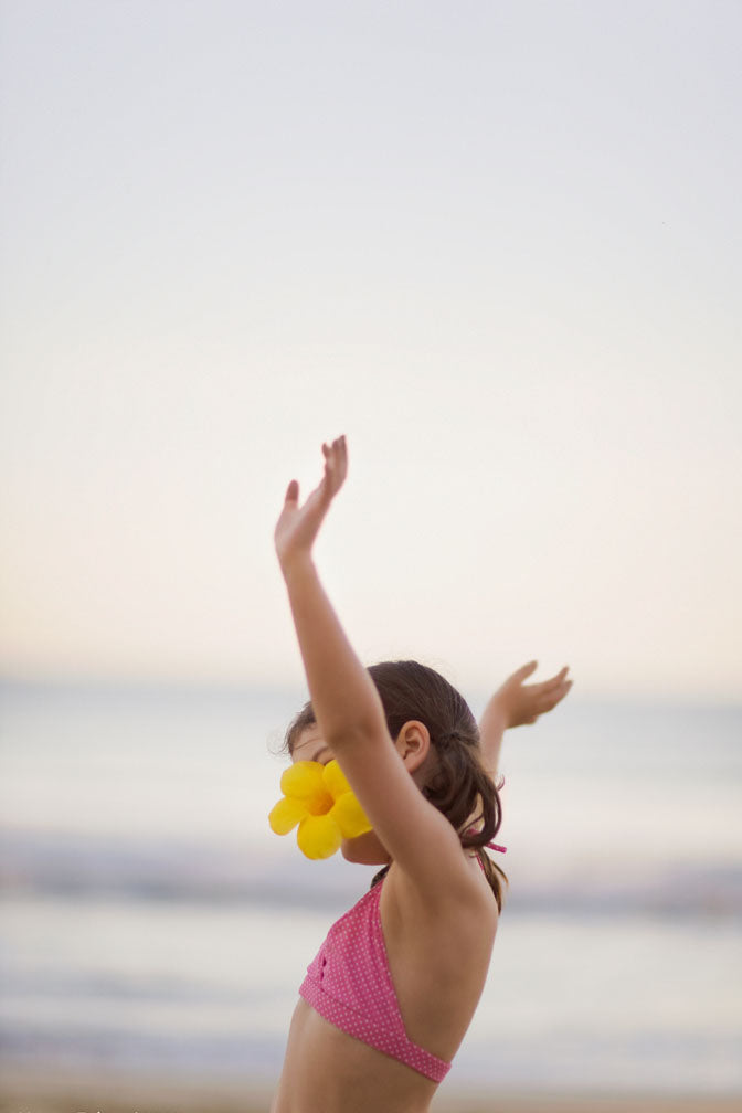 Child with a yellow flower in her hair standing on a beach with arms raised towards the sky.